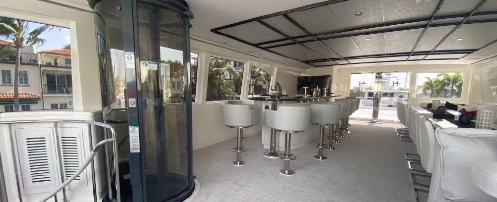 A modern interior of a yacht featuring a bar area with white stools, large windows, and a glass elevator.